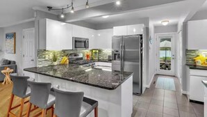 A modern kitchen with a granite island, stainless steel appliances, and a bar area with bar stools. - Beachside Bliss w/ Game Room, Gym & Golf Green (Westbrook)