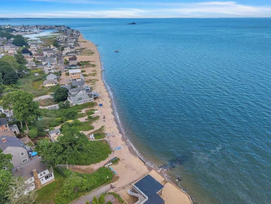 Vue aérienne d’une zone résidentielle côtière, avec des maisons le long de la plage, un ciel dégagé et une mer calme.
