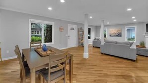 A spacious living room with a wooden dining table, gray sofa, and a large window. - Beachside Bliss w/ Game Room, Gym & Golf Green (Westbrook)