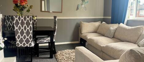 A living room with a beige sofa, a dining table with a patterned table runner, and a vase with flowers.