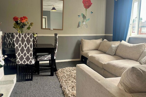 Ein Wohnzimmer mit einem beigen Sofa, einem Esstisch mit gemustertem Tischläufer und einer Vase mit Blumen.