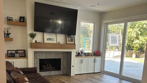 Amplia sala de estar con una gran televisión de pantalla plana sobre la chimenea, piso de madera y puerta corrediza de vidrio que da a un espacio exterior.