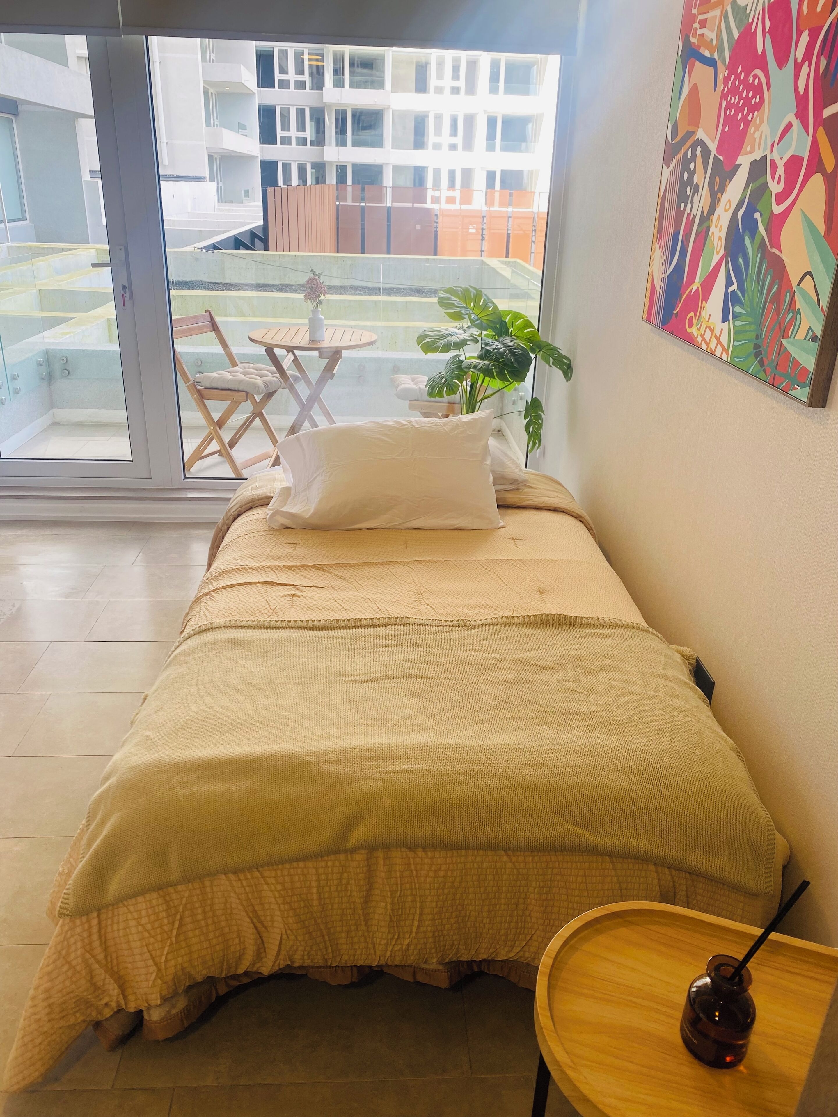 Une chambre avec un lit, une petite table en bois sur laquelle se trouve une boisson, une chaise et une vue sur un balcon agrémenté d’une plante et d’une table.