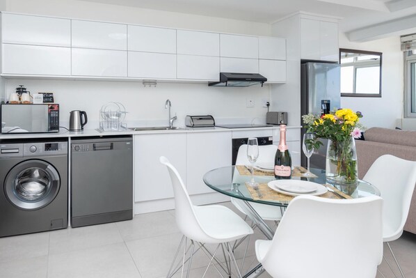 A modern kitchen with a dishwasher, washing machine, and a glass dining table with chairs. - Sea Views by the Promenade at Oceanmist Studio (Cape Town)