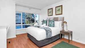 Un dormitorio moderno con una cama grande, una mesita de noche, un tapete verde y una ventana con vistas a un edificio y árboles.