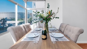 Mesa de comedor dispuesta para dos, con una botella de champán, un florero y vistas a un paisaje urbano a través de grandes ventanales.