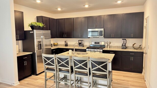A modern kitchen with dark cabinets, stainless steel appliances, a granite countertop, and a central island with bar stools. - Modern 4BR New Home w Luxury Touches (Del Valle)