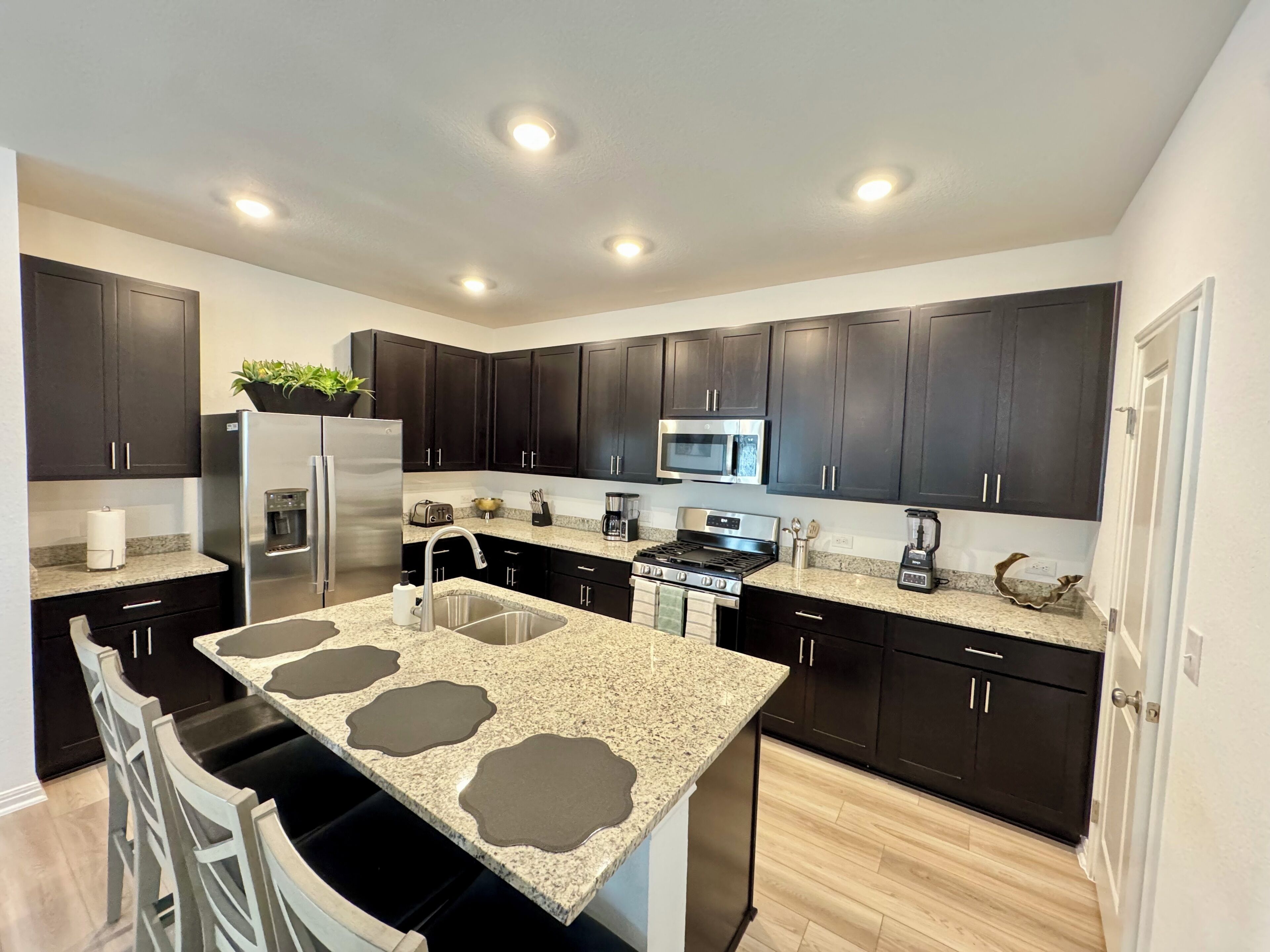A modern kitchen with dark cabinets, granite countertops, stainless steel appliances, and recessed ceiling lights.