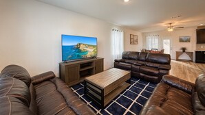 A living room with a large flat-screen TV, leather sofas, a wooden coffee table, and a patterned rug. - Modern 4BR New Home w Luxury Touches (Del Valle)