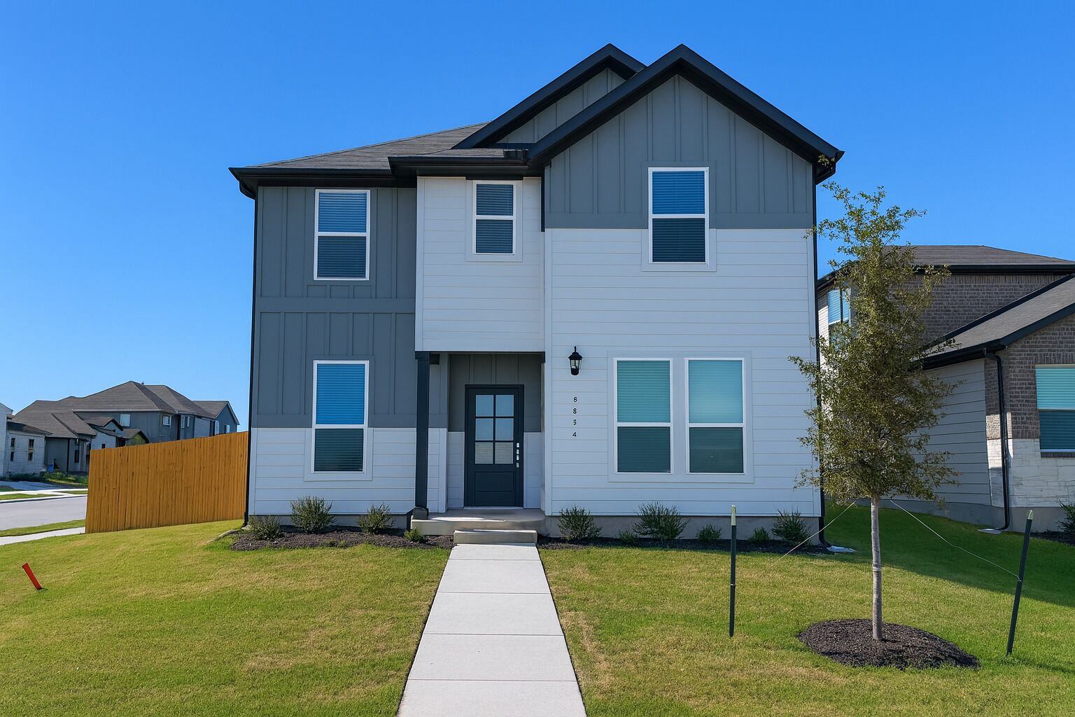 A two-story residential building with a grey and white exterior, a front door, and a small front yard with a tree and a wooden fence.