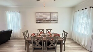 A dining area with a wooden table set for four, surrounded by chairs. A ceiling fan is above, and there are two windows with sheer curtains. - Modern 4BR New Home w Luxury Touches (Del Valle)