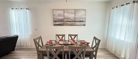 A dining area with a wooden table set for four, surrounded by chairs. A ceiling fan is above, and there are two windows with sheer curtains.