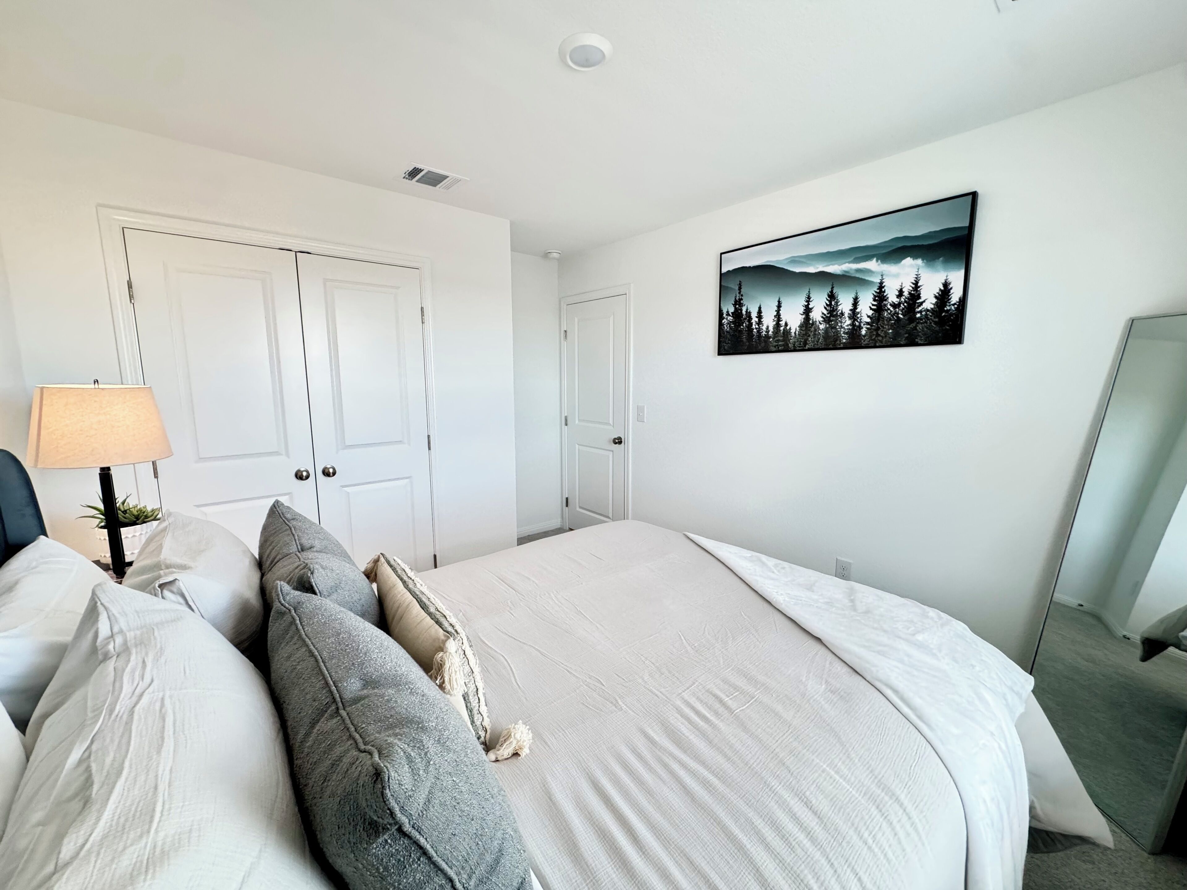 Una camera da letto con un letto, cuscini, una lampada, un quadro e uno specchio.