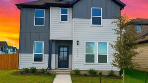 A two-story residential building with a grey and white exterior, a black front door, and a small front yard with a young tree and a sidewalk leading to the entrance. - Modern 4BR New Home w Luxury Touches (Del Valle)