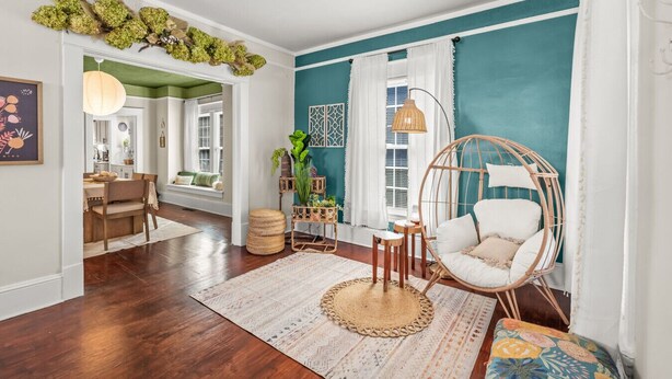 A room with teal walls, a round wicker chair, a woven rug, and a dining area with a wooden table and chairs.