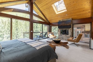 A spacious bedroom with a large bed, a stone fireplace, and a view of a forest through large windows.