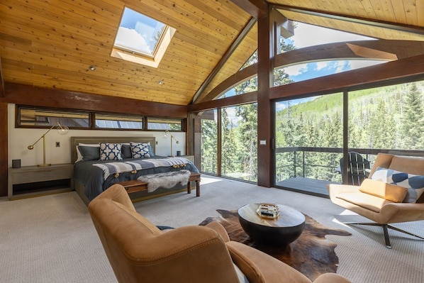 A spacious room with a large bed, a sofa, a coffee table, and a view of a forest through large windows.