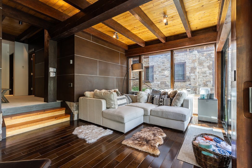 A modern living room with a sectional sofa, wooden flooring, a stone wall, and large windows.