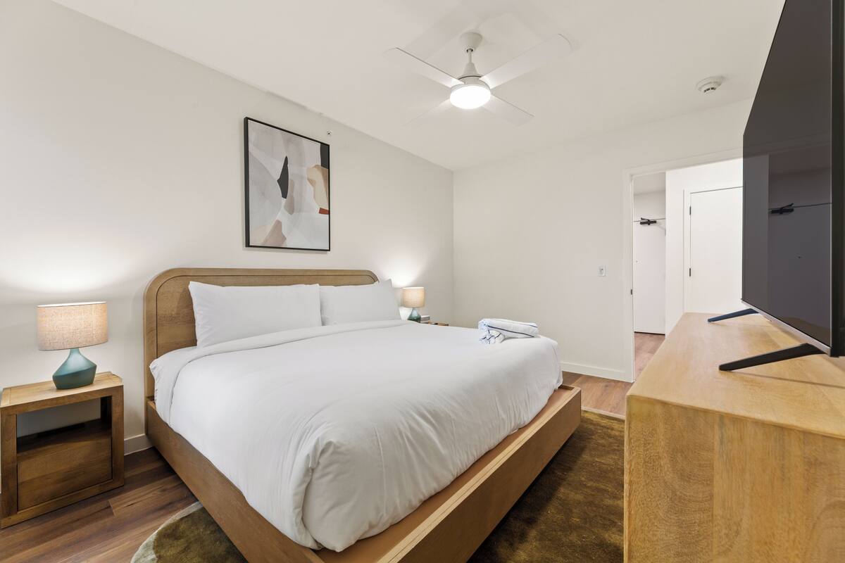 Un dormitorio con cama, mesitas de noche, ventilador de techo, televisor y una puerta.