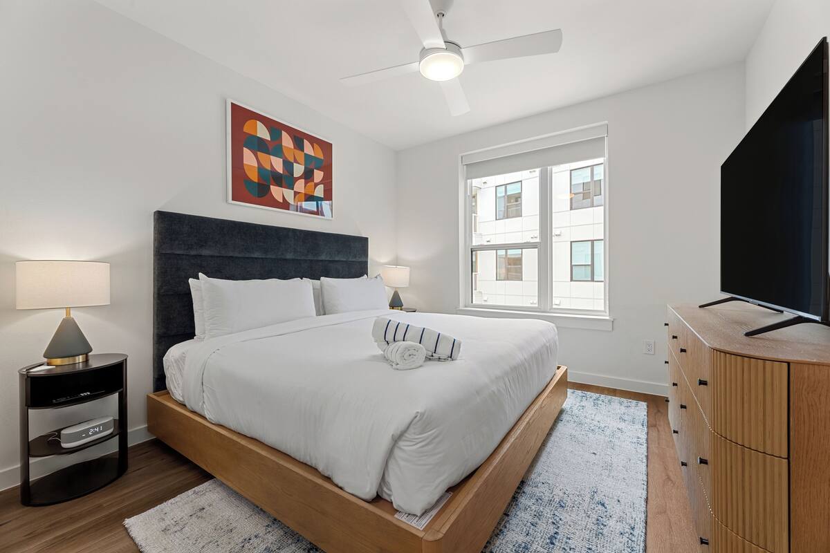 Un dormitorio moderno con cama, lámparas de noche, ventilador de techo, televisor y ventana con vista a edificios.