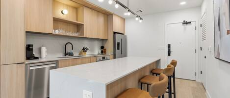 A modern kitchen with wooden cabinets, a stainless steel refrigerator, a white countertop, and bar stools.