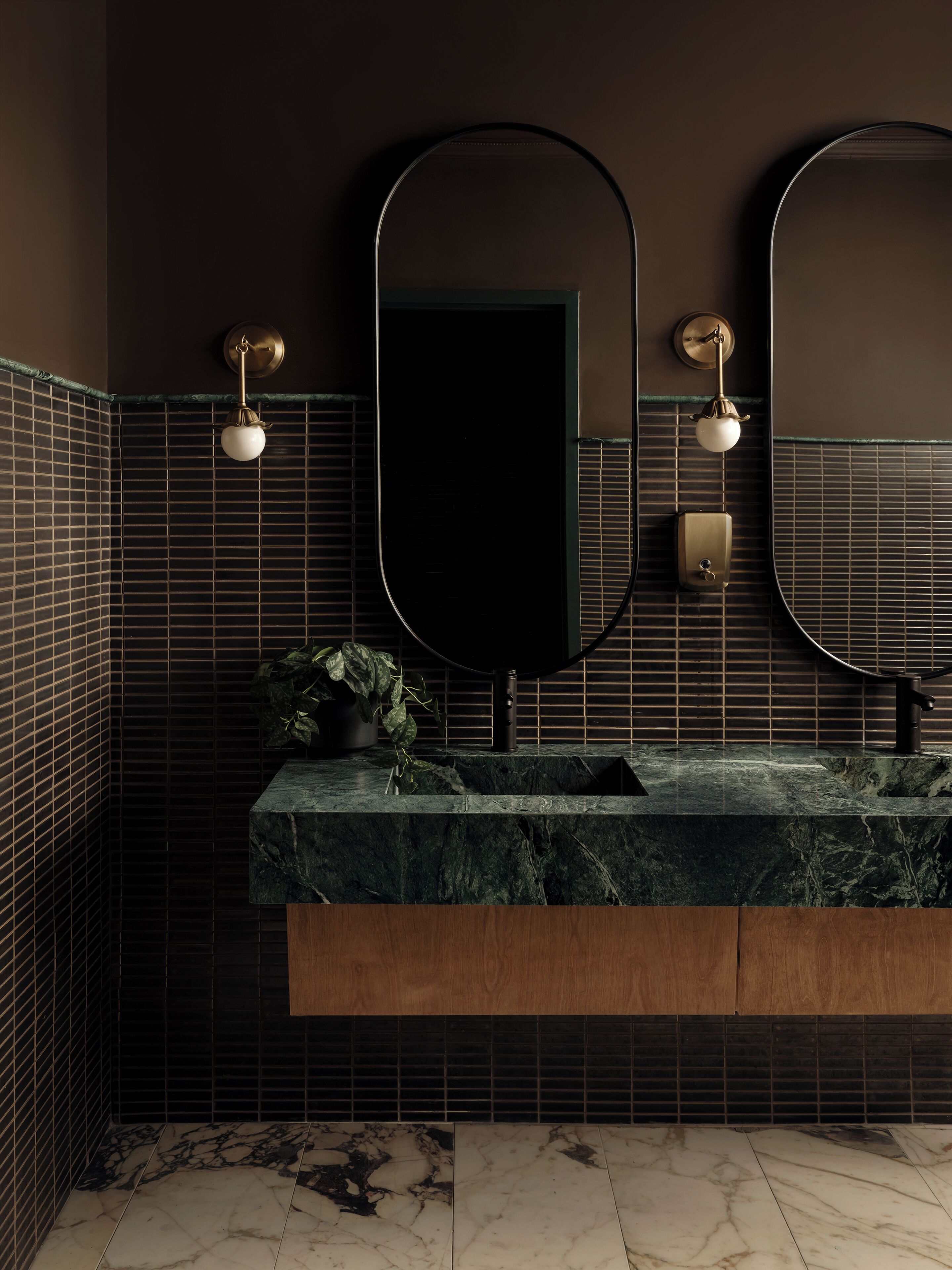 Un baño con pared de azulejos oscuros, un lavabo de mármol y dos espejos adosados a la pared.