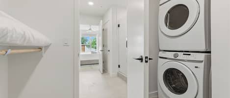 A laundry room with two stacked washing machines, a white door, and a hallway leading to a room with a bed and a window.