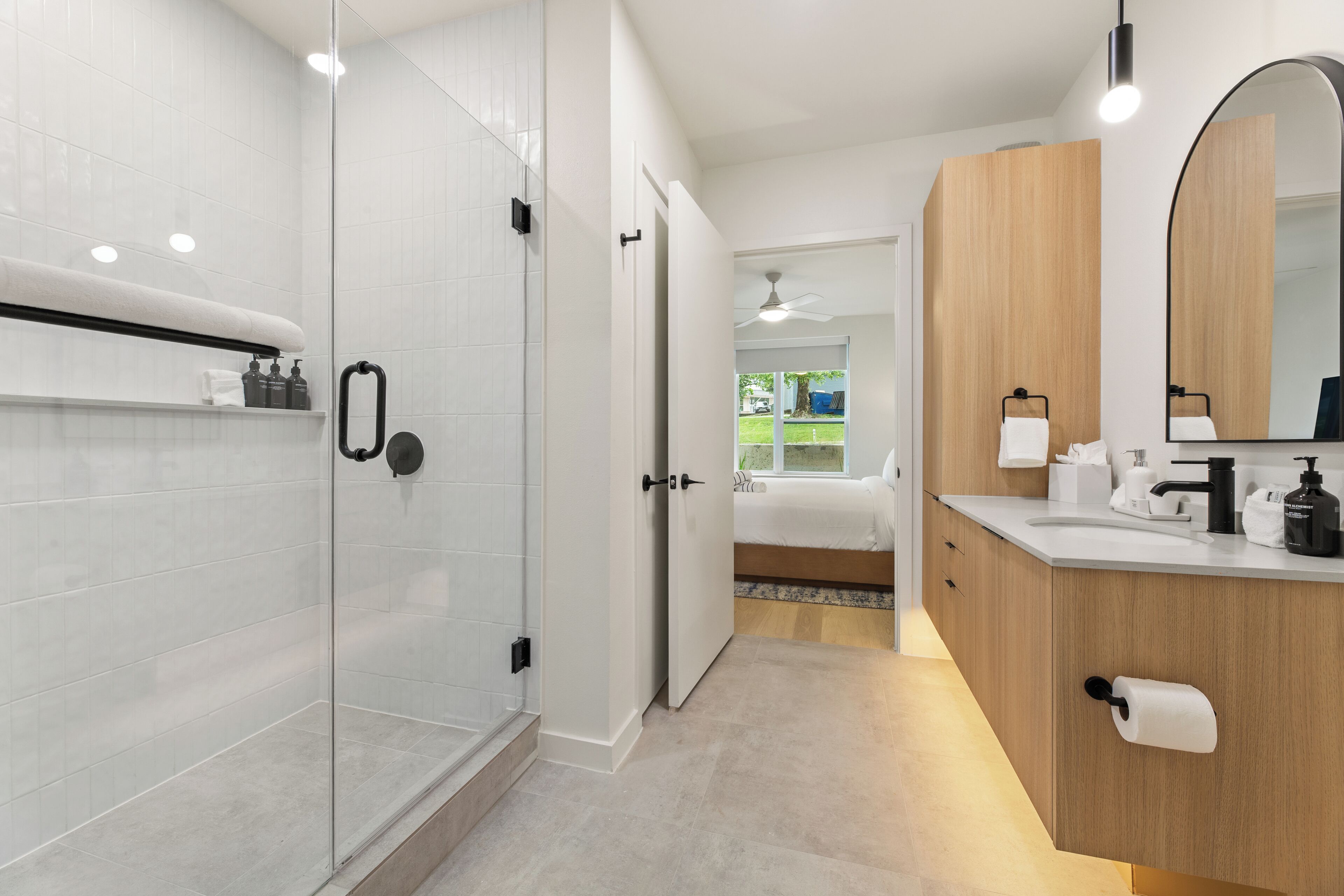 Une salle de bain moderne dotée d’une douche à parois de verre, d’un meuble-lavabo en bois avec un miroir, et d’une chambre à coucher visible avec un lit.
