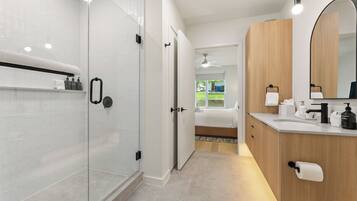 A modern bathroom with a glass shower enclosure, a wooden vanity with a mirror, and a visible bedroom with a bed.