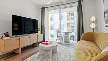 A modern living room with a flat-screen TV, wooden furniture, a round coffee table, a yellow sofa, and a large window with a view of a building.