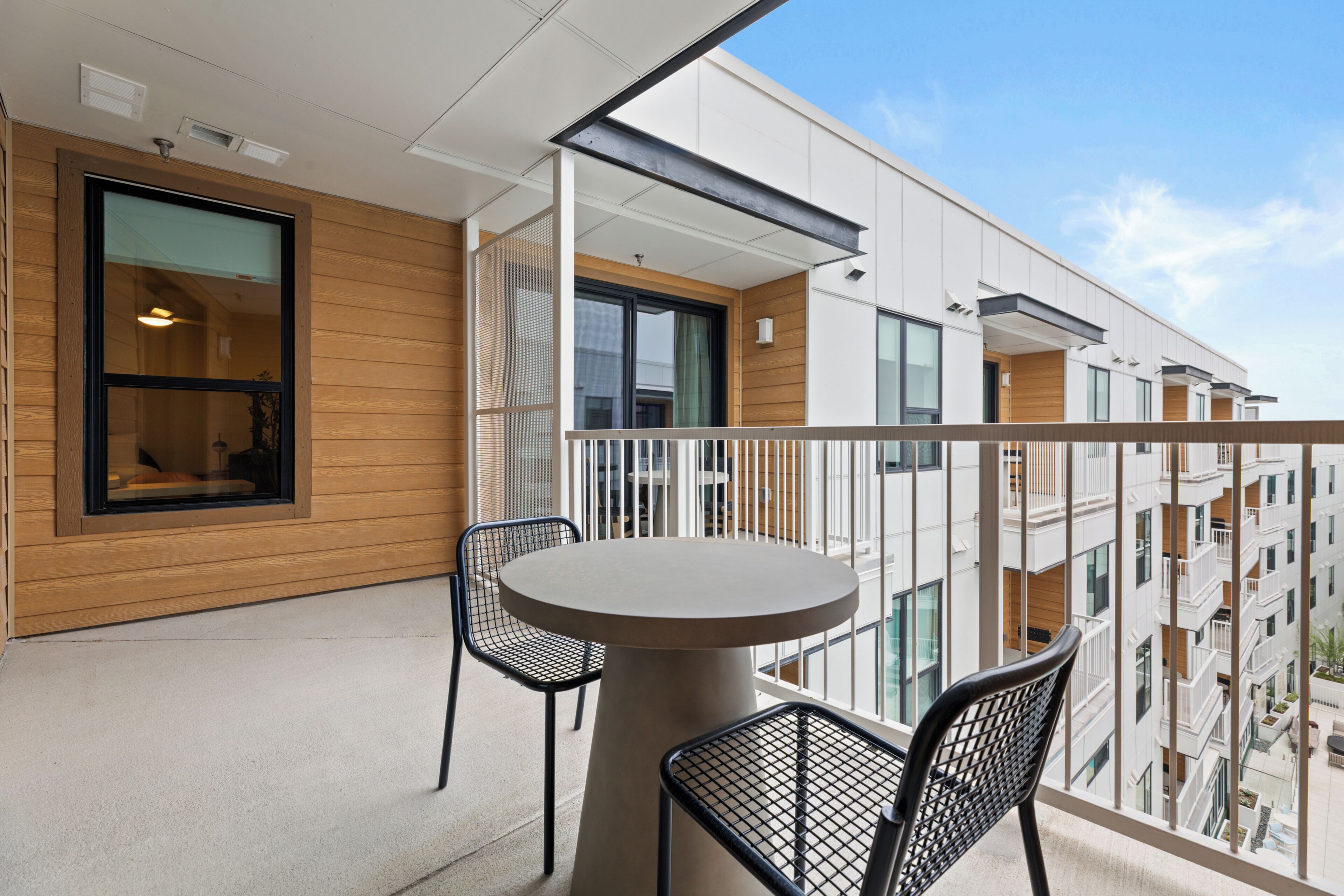 A balcony with a round table and two chairs, overlooking a modern building with multiple balconies.