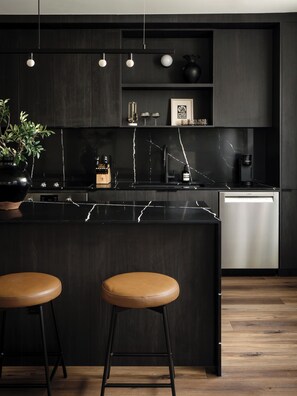 A modern kitchen with a marble countertop, two stools, a coffee machine, and a dishwasher.