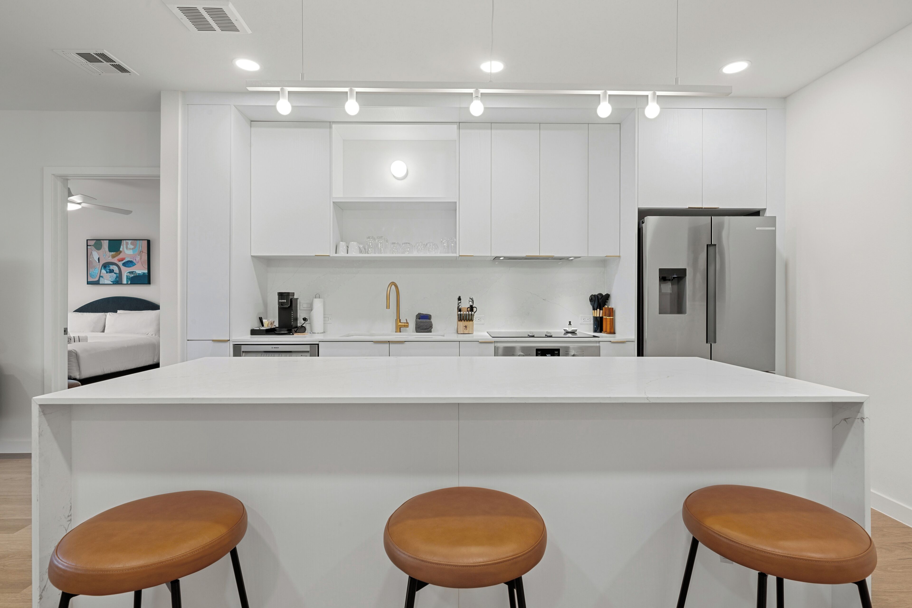 Cozinha moderna com armários brancos, uma ilha central com banquetas e uma geladeira de aço inoxidável.
