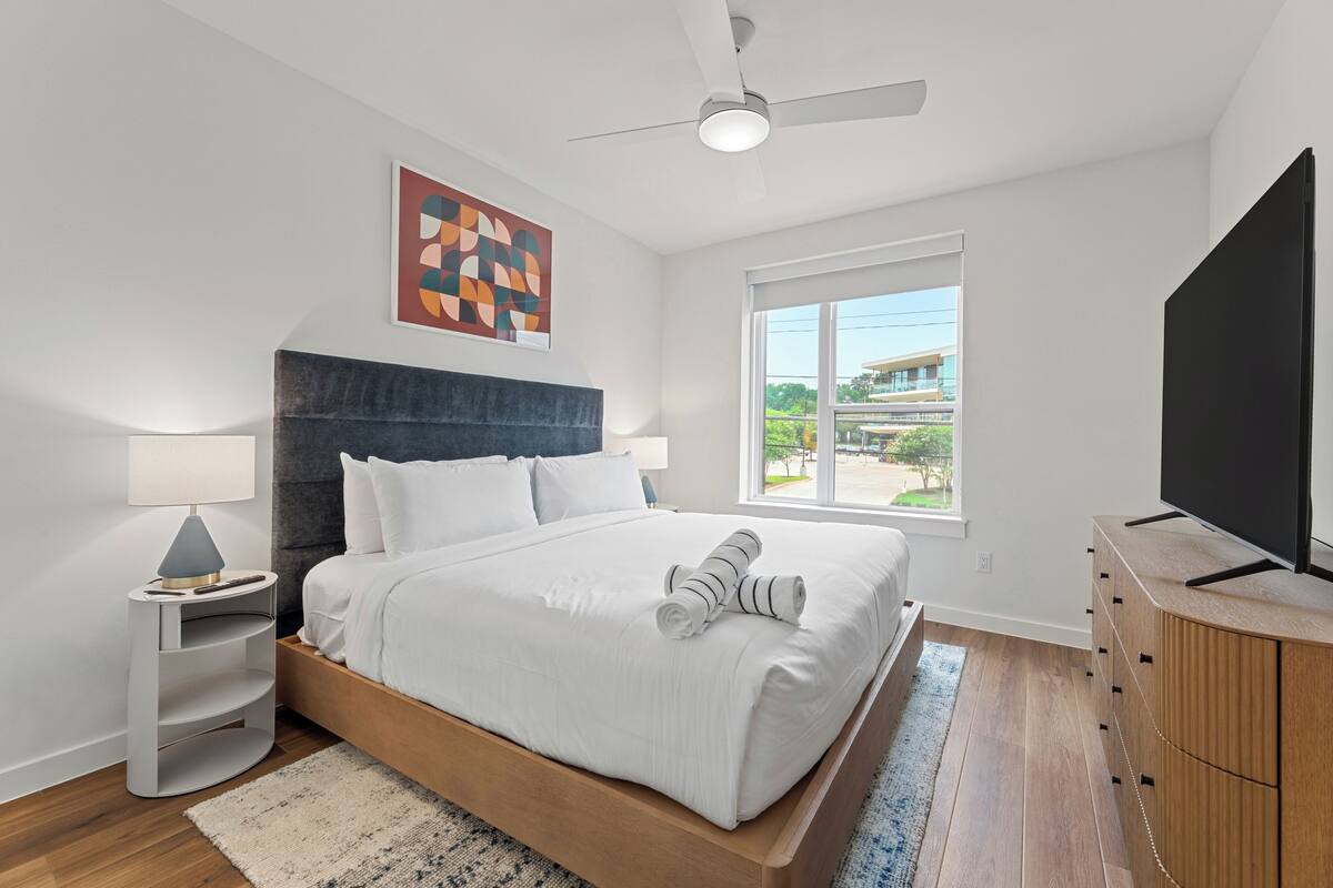 Un dormitorio moderno con cama, ventilador de techo, televisión y ventana con vistas.