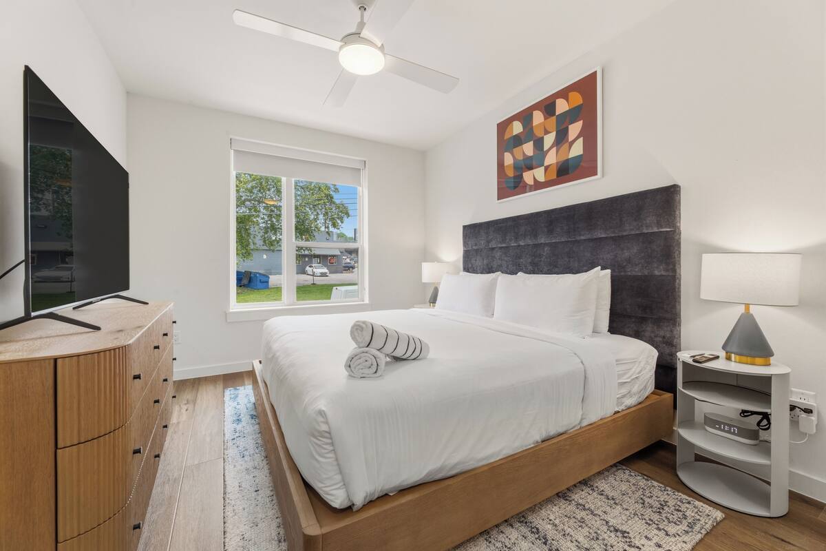 Un dormitorio con cama, televisión, ventilador de techo, ventana y un cuadro.