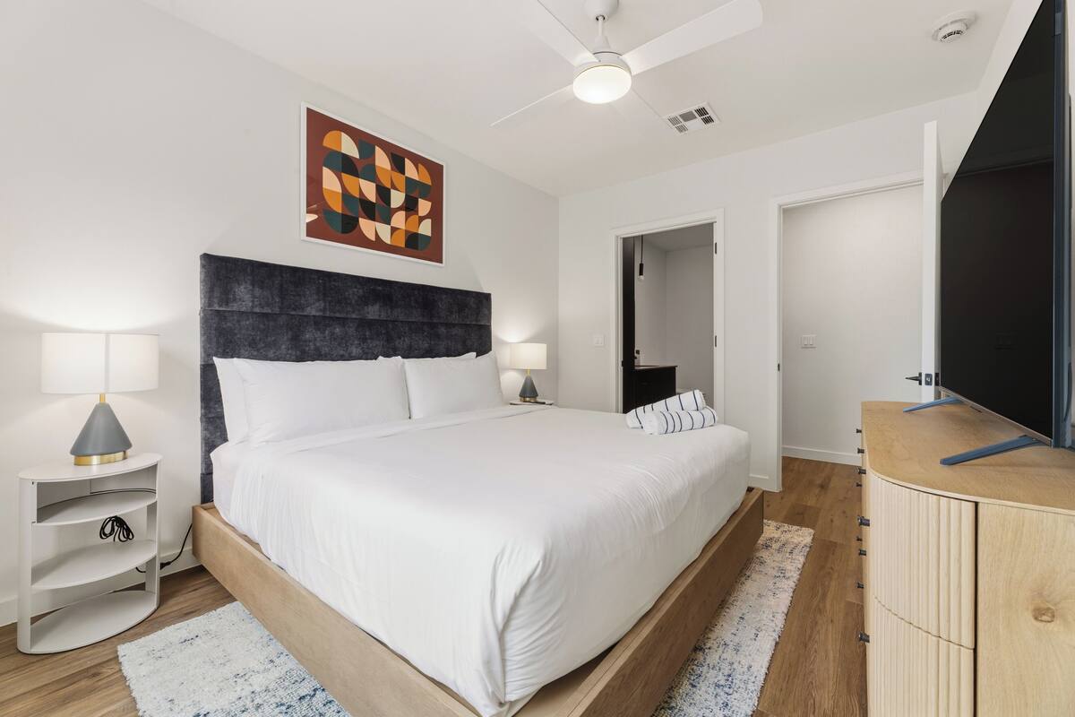 Un dormitorio moderno con cama, lámparas de noche, cabecera de madera, un mueble para televisor y ventilador de techo.