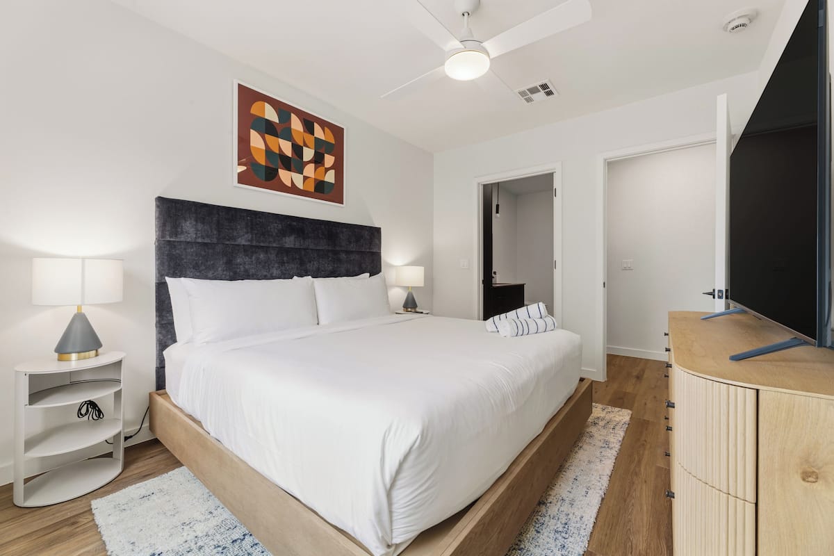 A modern bedroom with a bed, bedside lamps, a wooden headboard, a TV stand, and a ceiling fan.