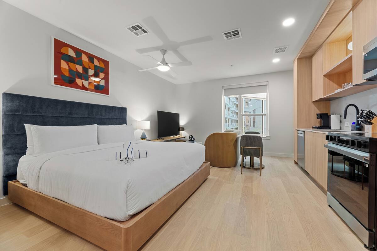 Habitación de hotel moderna con una cama grande, un escritorio, una silla, un televisor, ventilador de techo y una zona de cocina.