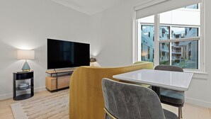 A modern living room with a flat-screen TV, a sofa, a dining table, and a large window with a view of buildings.