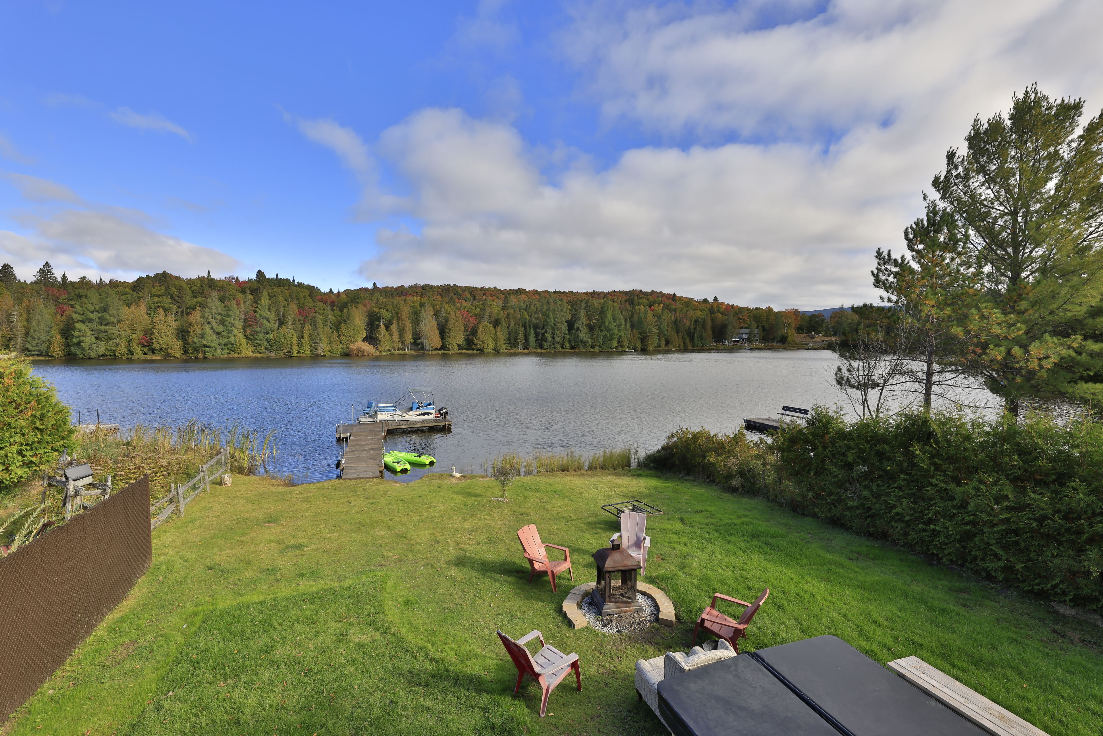 A lakeside patio with outdoor furniture, a dock with boats, and a forested backdrop.