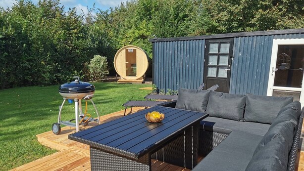 Eine Terrasse mit einem dunkelbraunen Tisch und einem Korbsessel mit grauen Kissen.