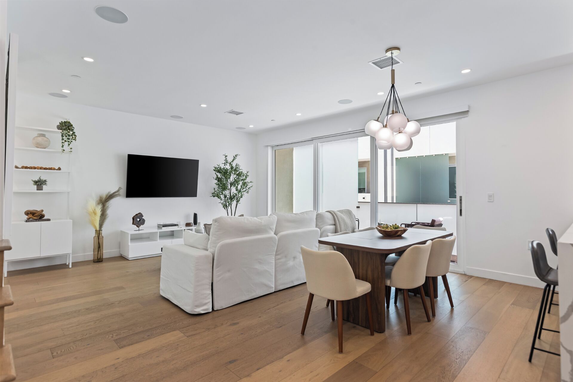 A modern living room with a white sofa, a dining table with chairs, a flat-screen TV mounted on the wall, and a large window providing natural light.