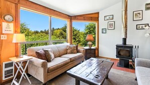 A cozy living room with a sofa, a table, a lamp, a fireplace, and large windows overlooking a forest. - Pet-friendly Oceanfront Getaway (Rockaway Beach)