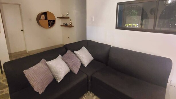 Ein Wohnzimmer mit einem schwarzen Ecksofa, einem runden Wandregal und einem Fenster mit Blick ins Freie.