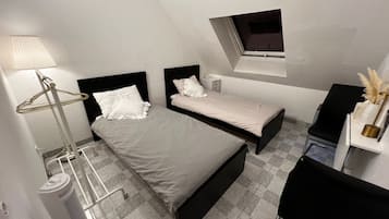 A bedroom with two beds, a window, a radiator, and a desk with a lamp and decorative items.