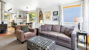 Een gezellige woonkamer met een bank, fauteuil, pouf en een open haard. Er is een eethoek met een tafel en stoelen, en een keuken op de achtergrond.