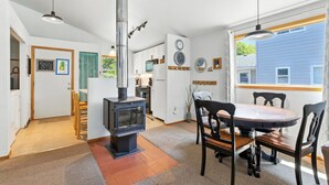 Een gezellige keuken met een houtkachel, een eettafel met stoelen en uitzicht op een blauw huis door het raam.