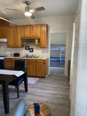 Une cuisine avec des meubles en bois, une table à manger avec des chaises, un ventilateur de plafond et une vue sur une chambre avec un lit superposé.