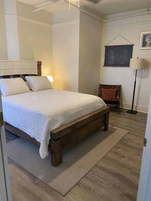Une chambre à coucher avec un lit en bois, une table de chevet, une chaise, une lampe sur pied et un tableau encadré au mur.