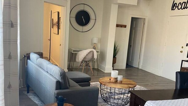 Ein modernes Wohnzimmer mit einer Couch, einem Couchtisch und einem Esstisch mit Stühlen. An der Wand hängt eine Uhr und es gibt einen Deckenventilator.